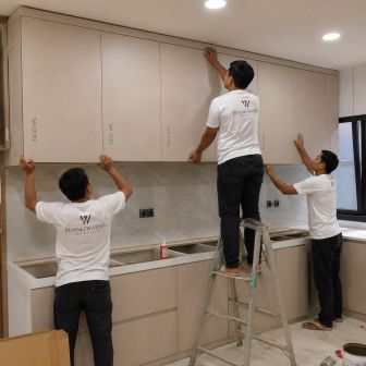 Tiga tukang Ruang Warna Interior Jakarta Timur sedang memasang lemari atas kitchen set compact menggunakan tangga dengan proses kerja yang rapi dan profesional.